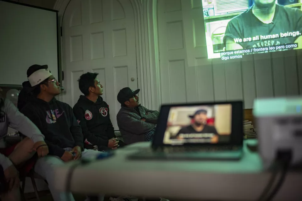 People sitting and watching a projected video with subtitles reading "We are all human beings," room decorated with balloons.