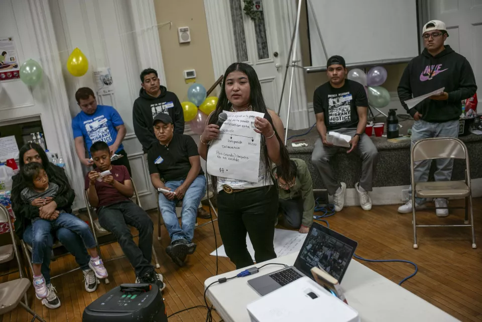 A young woman speaks into a microphone while holding handwritten notes, seated group behind her, community meeting.