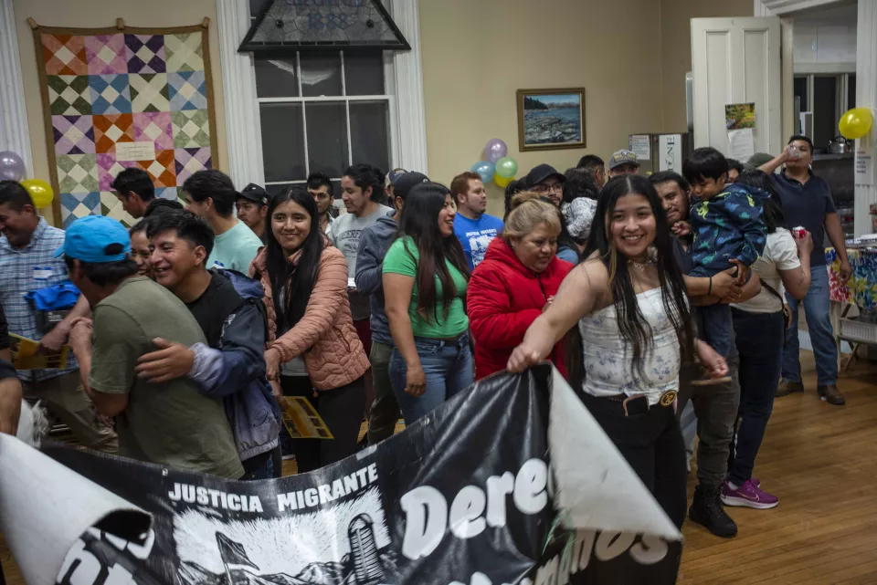 Crowd of people hugging and celebrating indoors, holding a "Justicia Migrante / Derechos" banner.