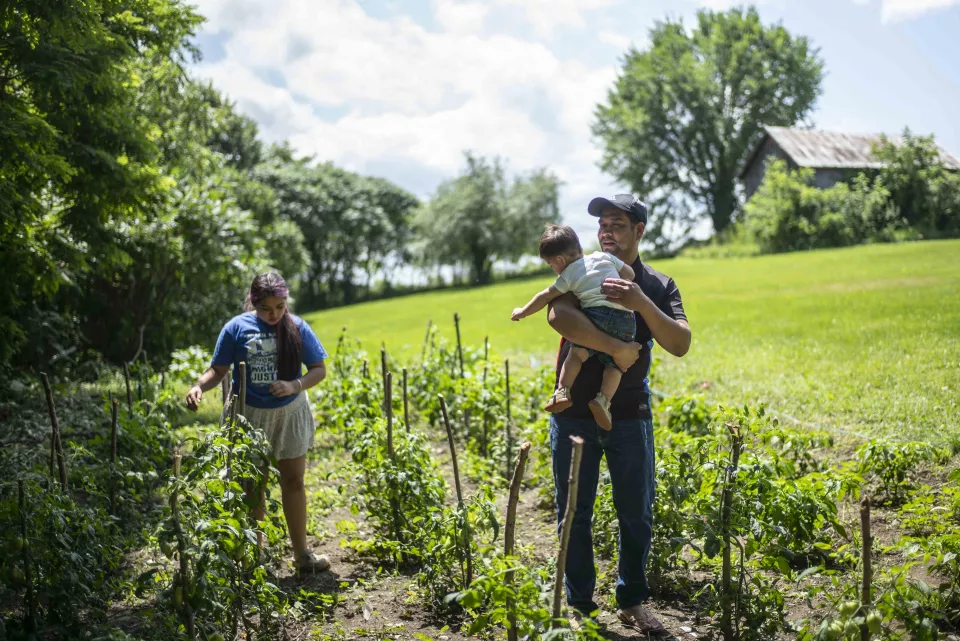 A man carrying a toddler walks through rows of a vegetable garden with a young woman, farm building in the background.