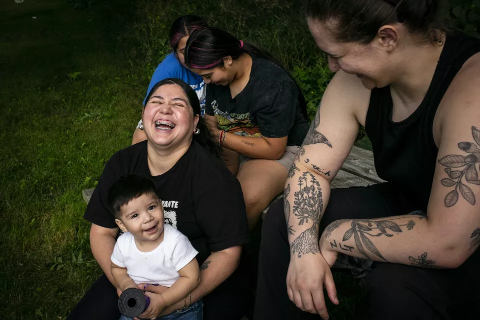 A woman laughs with a baby on her lap, other women sitting nearby outdoors, tattoos visible.