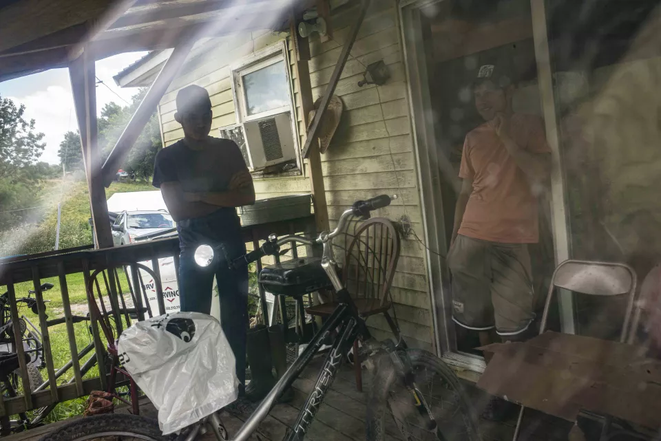 Two men on a porch, one leaning against a bicycle, reflection in a window.