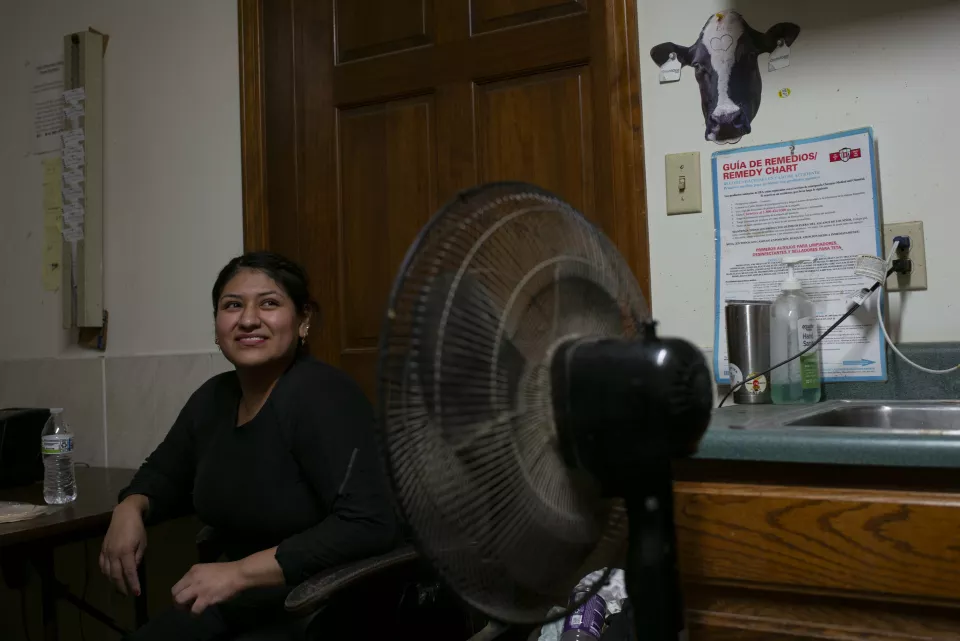 A smiling woman sits indoors near a fan, a "Gu&iacute;a de Remedios / Remedy Chart" poster and a cow photo on the wall behind her.