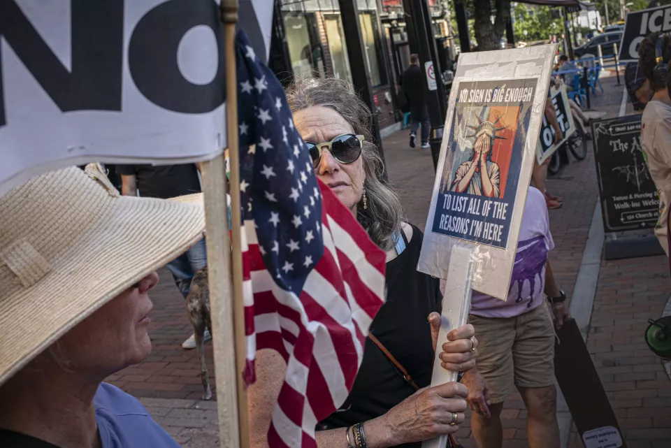 An older woman holds a sign reading "NO SIGN IS BIG ENOUGH TO LIST ALL OF THE REASONS I'M HERE," showing a weeping Statue of Liberty, an American flag beside her, another woman in a wide-brimmed sunhat in the foreground.