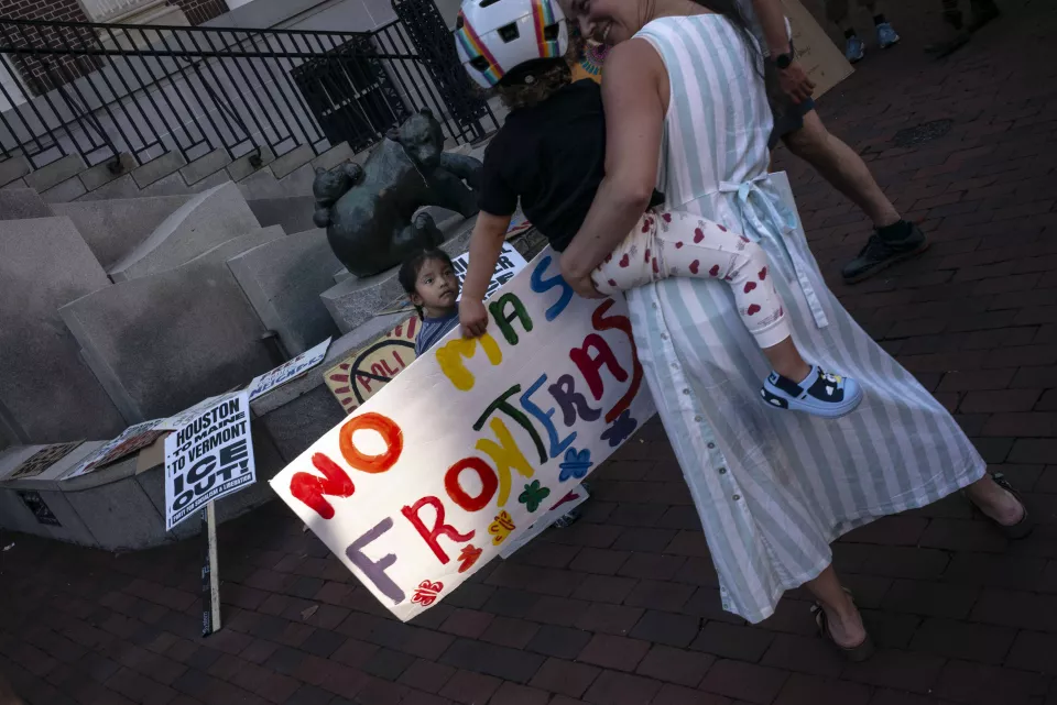 Overhead view of a woman carrying a child in a rainbow helmet down stone steps, a small girl standing near a bronze bear statue, a large hand-painted sign reading "NO MAS FRONTERAS" in the foreground.