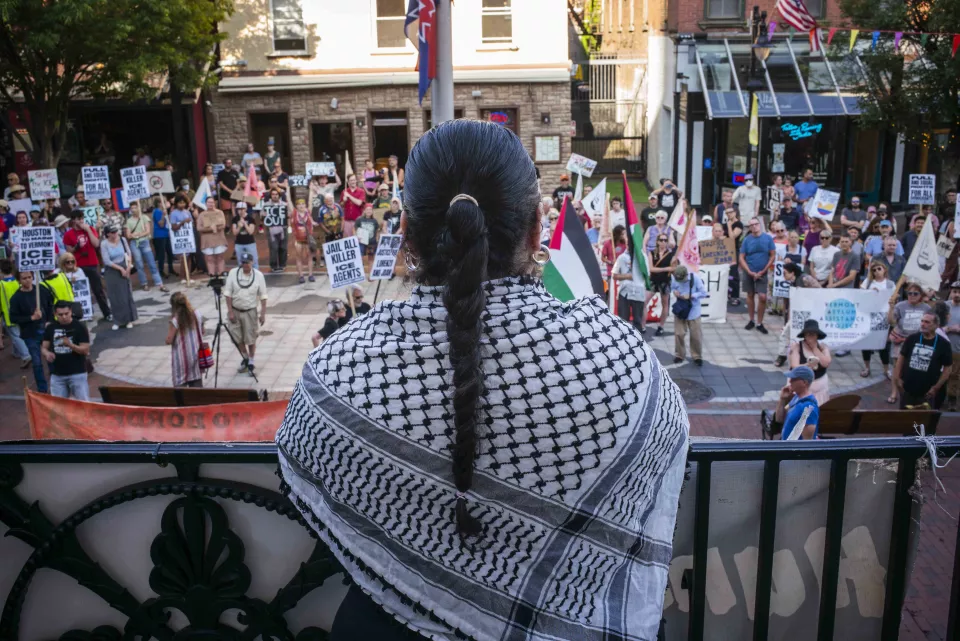 Back view of a woman wearing a black-and-white checkered keffiyeh, looking down from a balcony at a large rally crowd below holding signs and a Palestinian flag.