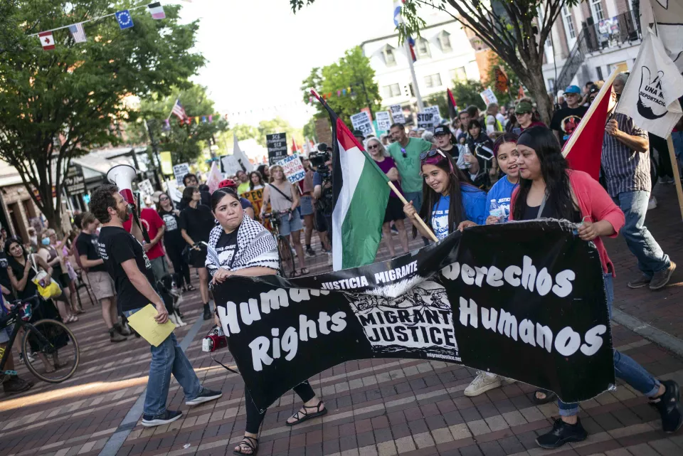 Street march with women carrying a banner reading "Human Rights / Derechos Humanos &ndash; Migrant Justice," a Palestinian flag waving above, a man with a megaphone nearby.