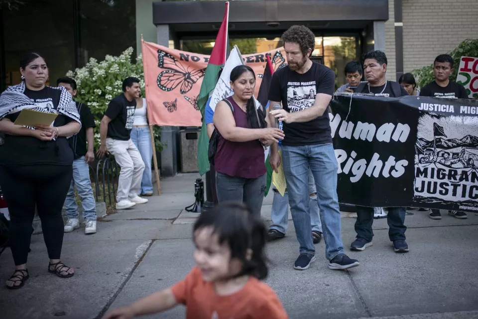 Rally outside a building, a man holds a microphone for a Spanish-speaking woman to address the crowd, a "Human Rights &ndash; Migrant Justice" banner and monarch butterfly flag behind them, a small child running in the foreground.