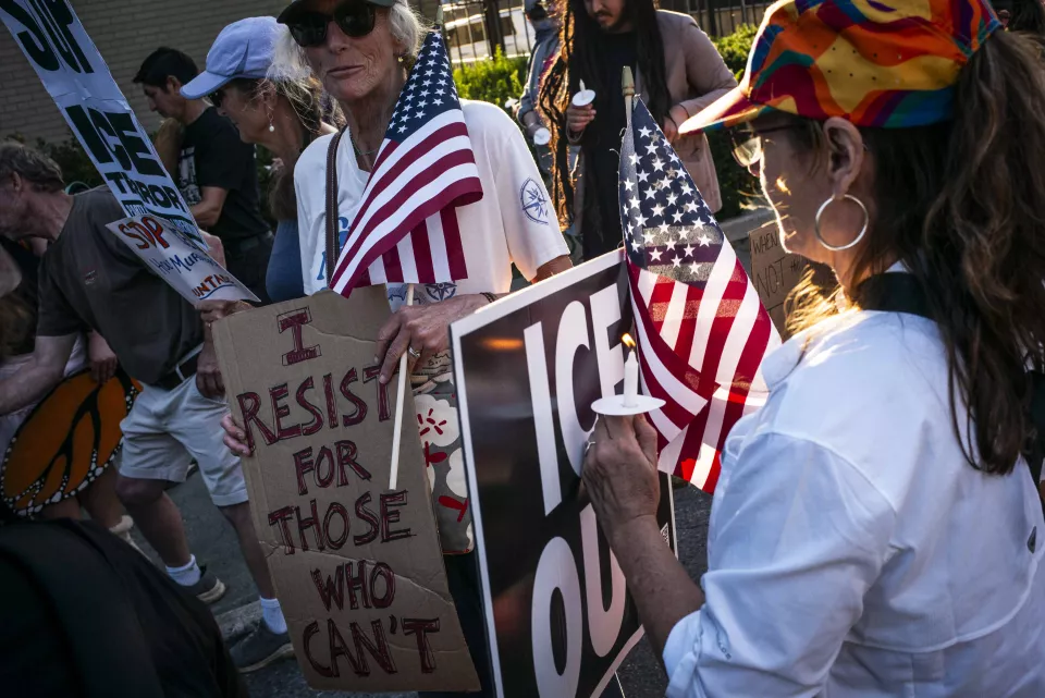 Close-up of a woman lighting a candle for another woman holding a sign reading "I RESIST FOR THOSE WHO CAN'T," American flags and an "ICE OUT" sign nearby.