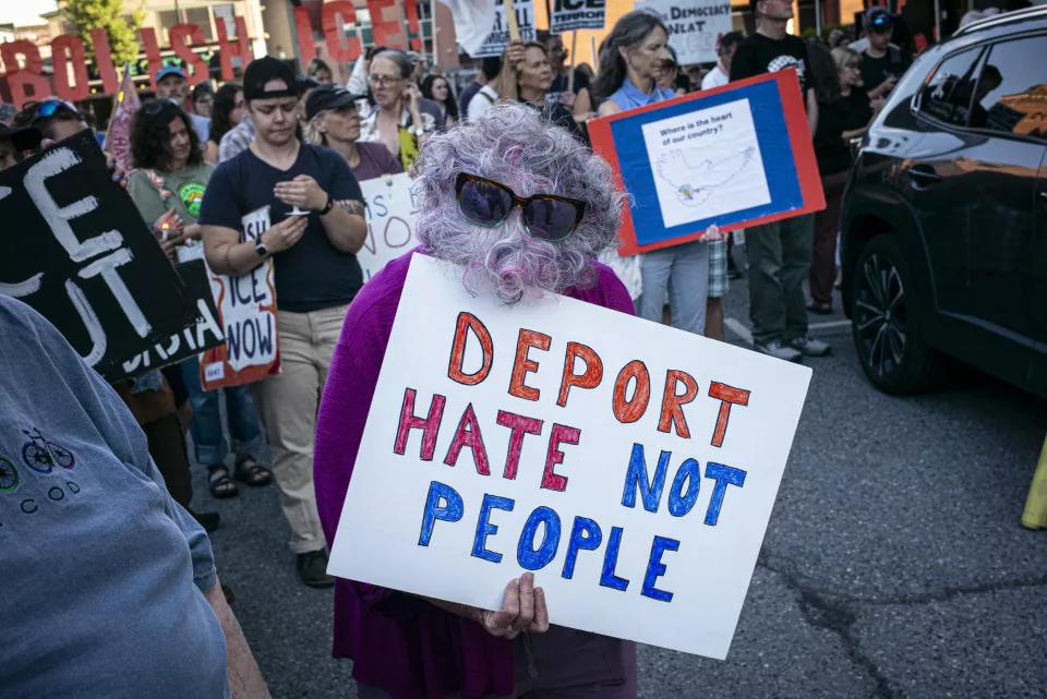 A woman in a purple top and curly gray-and-pink wig holds a sign reading "DEPORT HATE NOT PEOPLE," a crowd with more signs behind her.