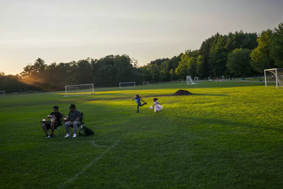 Family picnic in a park at sunset, people seated at a picnic table and in folding chairs near soccer goals, coolers and food spread on a table.